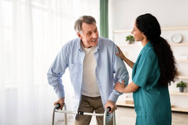 Genç hemşire huzurevinde yaşlı adamın çerçeveyle yürümesine yardım ediyor. Olgun erkek, minyon, bakıcı yaşlılar merkezinde geziyor. Profesyonel yaşlı bakım servisi.