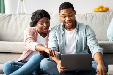 Şaşırmış Afro-Amerikan çift koltuğun yanında oturmuş dizüstü bilgisayar kullanıyor, sürpriz siyahi kadın kocasına veya erkek arkadaşına heyecan verici bir şey gösteriyor, defter ekranına işaret ediyor, boşluğu kopyalıyor.