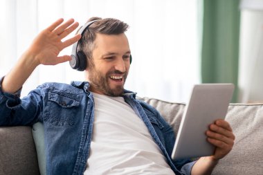 Neşeli orta yaşlı sakallı bir adam arkadaşlarıyla veya sevgilileriyle video görüşmesi yapıyor, dijital tablet ve kablosuz kulaklık kullanıyor, tablet ekrana bakıyor, gülümsüyor ve el sallıyor, ev içini kopyalıyor.