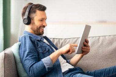 Kanepeye yaslanıp gülümseyen, iç mekan, yan görüş, fotokopi alanı, video izleyen veya müzik dinleyen ya da ped için mobil uygulama kullanan pozitif sakallı adam.