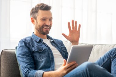 Mutlu yakışıklı bir adam arkadaşlarıyla ya da sevgilileriyle video konferansı yapıyor oturma odasında kanepeye yaslanıyor ve dijital tablet ekranına el sallıyor, boşluğu kopyalıyor. Kilitleme sırasında iletişim konsepti