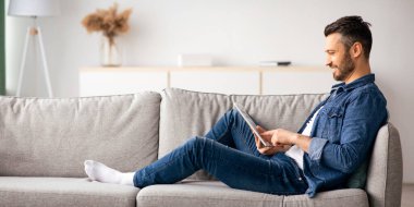 Oturma odasındaki kanepeye yaslanmış orta yaşlı bir adamın dijital tablet ve kopya alanı kullanarak yan görüntüsü. Mutlu sakallı adam evde eğleniyor, sosyal medyayı kontrol ediyor ya da arkadaşlarıyla sohbet ediyor.