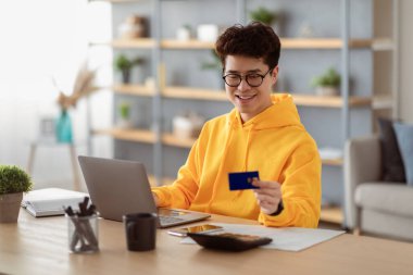 İnternetten alışveriş. Gözlüklü gülümseyen Asyalı bir adamın portresi PPC 'yi kullanarak masada oturuyor, kredi kartı alışverişine bakıyor, evdeki internet dükkanından para transfer ediyor. Omni Kanalı