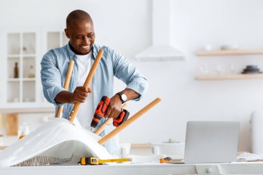 Çevrimiçi ders ve mobilya montajı için talimatlar. Yakışıklı Afrikalı Amerikalı adam elektrikli tornavidayla sandalye topluyor ve mutfaktaki dizüstü bilgisayara bakıyor.