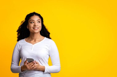 Ellerinde akıllı telefon tutan ve uzaklara bakan dalgın genç siyahi kadın, hayali Afrikalı Amerikalı kadın mobil teklif hakkında düşünüyor, sarı arka planda tek başına duruyor, kopyalama alanı olan panorama