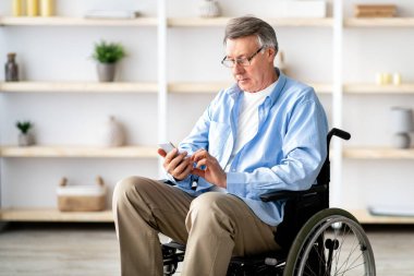 Tekerlekli sandalyedeki yaşlı adam akıllı telefon kullanıyor, mesajları kontrol ediyor, video izliyor, internette geziniyor. Modern mobil cihaz kullanan olgun engelli erkek.