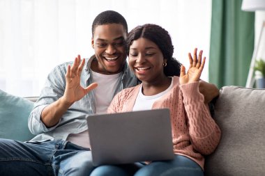 Neşeli siyah çift dizüstü bilgisayara el sallıyor, arkadaşları ya da akrabalarıyla görüntülü görüşme yapıyorlar, ev içi. Afrikalı Amerikalı genç aşıklar aileleriyle çevrimiçi parti yapıyor, gülümsüyor, kopyalama alanı