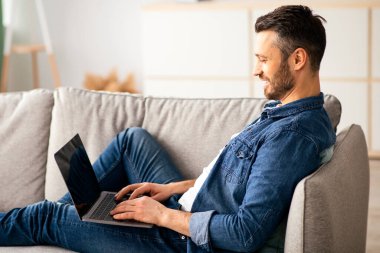 Oturma odasındaki koltukta dinlenen orta yaşlı bir adamın yan görüntüsü, en yeni dizüstü bilgisayarını kullanarak, kopyalama alanı. Mutlu sakallı adam evde eğleniyor, sosyal medyayı kontrol ediyor ya da bilgisayardaki arkadaşlarıyla sohbet ediyor.