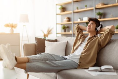Dinlen ve rahatla kavramı. Sakin Asyalı bir adam kanepede oturup müzik, ses kitabı, podcast dinliyor, uyku için meditasyonun tadını çıkarıyor ve kablosuz kulaklıklarla huzur dolu bir zihin, arkasına yaslanıyor, uzayı kopyalıyor.