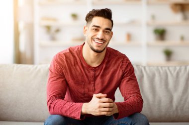 Yakışıklı Arap adam evde takılıyor, kanepede oturuyor ve kameraya gülümsüyor, fotokopi çekiyor. Neşeli bir orta doğulu gencin portresi. Hafta sonunun tadını çıkarıyor. Oturma odasındaki kanepede dinleniyor.