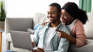 Neşeli Afro-Amerikan erkek ve kadın evde otururken web sörfü yapıyor, oturma odasında dizüstü bilgisayar kullanıyor. Not defterine işaret eden mutlu siyah kadın, online alışveriş yapan genç çift, kopyalama alanı