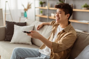 Boş zaman ve eğlence. Elinde uzaktan kumandayla televizyon ya da film izleyen gülümseyen Asyalı adam. Genç adam evdeki rahat kanepede oturup boş zamanın tadını çıkarıyor.