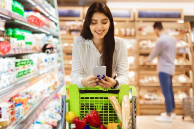 Süpermarkette akıllı telefon kullanan mutlu Orta Doğulu kadın markette alışveriş yapıyor ve kapalı alanda alışveriş arabasıyla yürüyor. Telefonunuz için market alışverişi cep telefonu uygulaması. Seçici Odaklanma