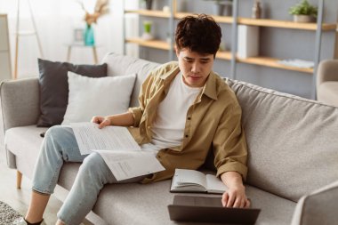 Evrak işi kavramı. Evde çalışan Asyalı bir gencin portresi, elinde kağıt ya da finansal belge tutarken, kanepede otururken ve oturma odasında dizüstü bilgisayar kullanırken, klavyede yazarken.