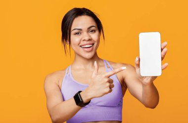Spor uygulaması konsepti. Afro-Amerikan kadını elinde tuttuğu ve siyah ekranlı akıllı telefonu işaret ettiği için mutlu. Sarı arka planda duruyor.