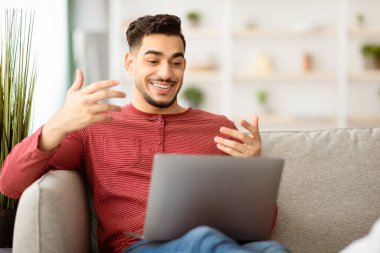 Mutlu Müslüman adam dizüstü bilgisayarla parti veriyor oturma odasında kanepede oturuyor, fotokopi çekiyor. Neşeli Arap adam evden video görüşmesi yapıyor, modern bilgisayar kullanıyor, iş görüşmesi yapıyor.