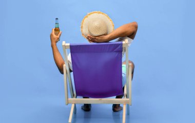 Genç siyahi adamın şezlongda oturmuş bira şişesiyle yaz tatilinin tadını çıkarırken, alkollü içecek yudumlarken, mavi stüdyo arka planında güneşlenirken,