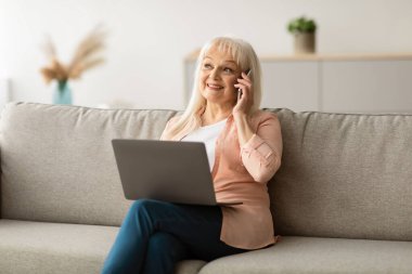 Mobil İletişim. Olgun bir kadının akıllı telefonuyla konuşması, evde dizüstü bilgisayarla çalışması, yaşlı bir kadının bilgisayar kullanması, hoş bir telefon konuşması, koltukta oturması...