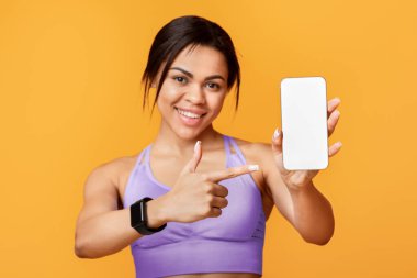 Spor başvurusu. Akıllı telefonu tutan ve gösteren heyecanlı siyahi kadın, model şablonu için siyah boş ekrana işaret ediyor, fitness takip cihazı takıyor, sarı stüdyo geçmişi var.