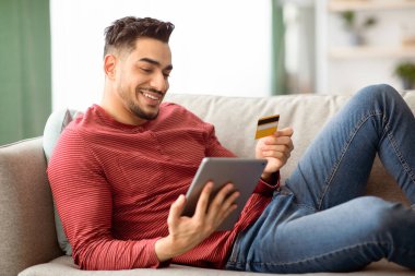 Evdeki kanepede dinlenirken, kredi kartı ve dijital tabletle neşeli genç bir Arap. İnternetten alışveriş yapan mutlu bir orta doğulu adam. Blok, e-ticaret, yan görüş kullanıyor.