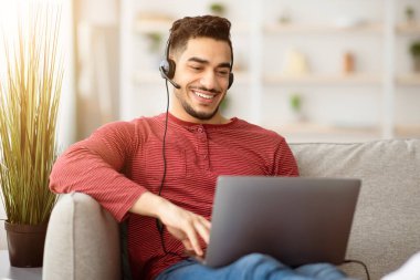 Mutlu Arap adam evde otururken internete bağlanıyor, dizüstü bilgisayar ve kulaklık kullanıyor, bilgisayar klavyesinde daktilo kullanıyor, oturma odasında kanepede oturuyor, boşluğu kopyalıyor. E-eğitim, çevrimiçi kurs kavramı