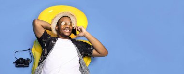 Havalı yaz kıyafetleri içinde havalı siyah bir adam şişme liloya uzanmış mavi stüdyo arka planında akıllı telefonla konuşuyor. Afrika kökenli Amerikalı bir adam plaj tatilinde telefonla konuşuyor.