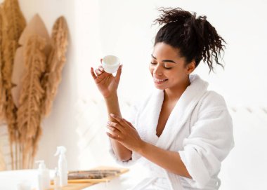 Güzel siyah kadın nemlendirici dirsekler nemlendirici krem sürerken kuru cilt üzerine modern banyoda küvette oturuyor. Dişi Güzellik ve Cilt Rutini Konsepti