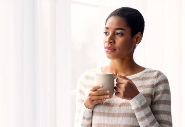 Genç siyahi kadın, evdeki pencerenin yanında dururken kahve içiyor, Milenyum Afrikalı Afrikalı Amerikalı kadının elinde fincanı tutuyor, sıcak içkinin ve şehrin manzarasının tadını çıkarıyor,