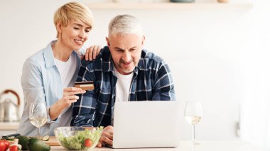 Ortak alışverişler, çevrimiçi mağazalar, salgın sırasında teslimat servisi. Mutlu yetişkin kadın ve erkek dizüstü bilgisayara bakar ve mutfak içinde kredi kartı tutarlar. Masada parlak sebzeler ve şarap bardakları vardır.