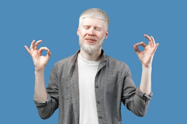 İyi iş çıkardın. Heyecanlı albino adam iki eliyle ok işaretleri yapıyor, iyi olduğunu gösteriyor, mavi stüdyo arka planında duruyor. Olağanüstü görünüşlü, soluk tenli ve beyaz saçlı genç bir adam.