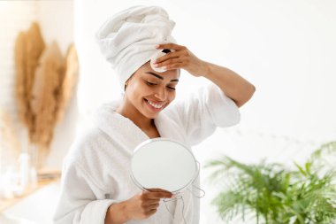 Genç siyahi kadın pamuk pedle makyaj yapıyor. Yuvarlak aynaya bakıyor. Modern banyoda duruyor. Yüz Bakımı, Güzellik ve Yüz Bakımı Konsepti