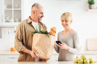 Covid-19 tecrit sırasındaki ev yemeği dağıtım uygulamasını inceleyin ve değerlendirin. Çevre çantalı mutlu yetişkin bir adam, market alışverişi yapıyor, karısı mesajı okuyor ve modern mutfağın içindeki servisi değerlendiriyor.