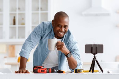 Mobilyaların montajı için video talimatları. Bir fincan kahveyle mutlu siyah bir adam web kamerasında akıllı telefona bakıyor, mutfak içinde masada aletlerle, boş bir alanda.