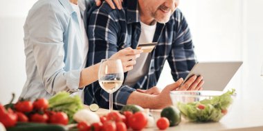 Çevrimiçi hizmet, COVid-19 karantinası sırasında alışveriş. Gülümseyen yetişkin kadın ve erkek tablete bakar ve mutfak içinde kredi kartı tutarlar. Masada parlak sebzeler, bardaklarda şarap, boş alan.