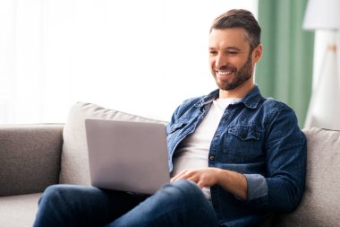 Modern dizüstü bilgisayar kullanan, yarı zamanlı çalışan, orta yaşlı gülümseyen bir adam. Mutlu sakallı adam film izliyor ya da dizüstü bilgisayarda sohbet ediyor oturma odasındaki kanepede dinleniyor.