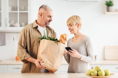 COVID-19 salgını sırasında aile için ev yapımı yemek, alışveriş ve sipariş teslimatı için ürünler. Elinde yiyecek dolu kağıt torbalarla gülümseyen son sınıf öğrencisi. Telefonlu kadın mutfaktaki pakete bakıyor.