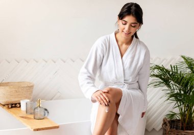 Banyodan çıktıktan sonra banyo küvetinde duran nemlendirici, pürüzsüz bacakları ve vücut losyonuyla kendini önemseyen çekici bayan. Cilt Bakımı ve Şımartma, Kadın Güzellik Ritüelleri
