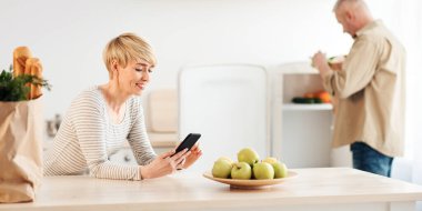 Yemek siparişi vermek, salgın sırasında eve erzak götürmek için bir uygulama. Eko torbalı mutlu orta yaşlı erkek, buzdolabına sebze atıyor, karısı modern mutfak içinde mesaj okuyor, boşluğu kopyalıyor.