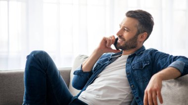 İletişim konsepti. Rahat orta yaşlı bir adam oturma odasında koltukta oturuyor, cep telefonuyla arkadaşlarıyla veya sevgilileriyle konuşuyor ve fotokopi odasına bakıyor.
