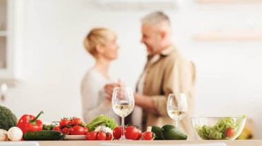 Covid-19 tecrit, romantik randevu, aile ilişkileri. Minimalist beyaz mutfak içinde dans eden mutlu yaşlı çift. Şaraplı bardaklara odaklanın ve parlak sebzeler ve masada salata, bulanık