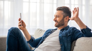Yakışıklı bir adamın arkadaşlarıyla ya da sevgilileriyle telefon aracılığıyla video görüşmesi yaparken oturma odasında koltuğa yaslanırken ve cep telefonu ekranına el sallarken yan görüntüsü. Kilitleme sırasında iletişim konsepti