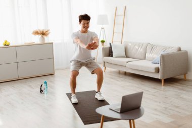 Yurtiçi antreman. Gülümseyen Atletik Asyalı Adam evde çalışırken diz üstü bilgisayar ekranına bakarak çömeliyor. Erkek Fitness Rutini. Sportif Adam Bilgisayar Seçici Odaklanma Önünde Egzersiz Yapıyor