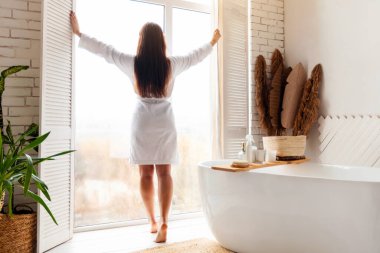 Sabah Güzelliği Rutini. Banyoda pencereleri açan, kameraya geri dönen, beyaz bornoz giyen, evdeki modern banyoda kaplıca ayinlerine hazırlanan bir kadın..