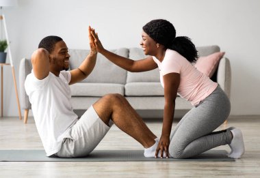 Atletik siyah çift evde birlikte egzersiz yapıyor, karın kasları üzerinde çalışıyor. Neşeli Afro-Amerikan kadın spor kıyafetleri içinde erkek bacaklarını tutuyor göbek egzersizleri yapıyor, tezahürat yapıyor.