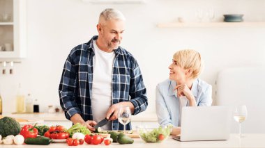 Sağlıklı yemek, romantik yemek ve birlikte yemek pişirmek. Neşeli orta yaşlı eş yemek tarifi için dizüstü bilgisayar kullanıyor ve yaşlı kocaya bakıyor, modern aydınlatma mutfağında sebze kesiyor, boş alan arıyor.