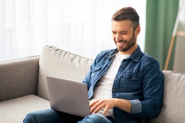 Gülümseyen orta yaşlı blog yazarı kanepe oturma odasında oturuyor, modern laptop kullanıyor, klavyeye yazıyor, makale yazıyor ya da takipçilerle sohbet ediyor. Mutlu adam evden çalışıyor, boşluğu kopyalıyor.