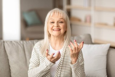 Çevrimiçi Video Çağrısı. Kameraya bakan, konuşan, arkadaşlarıyla sanal bir konuşma yapan, evdeki kanepede oturan olgun bir kadının portresi.