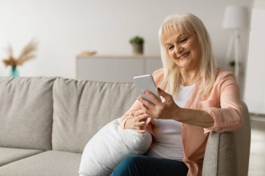 Ev eğlencesi ve boş zaman. Olgun bir bayanın elinde akıllı telefon tutması, mesaj yazması, koltukta oturması. İletişim Konsepti. İnternet 'te sörf, sohbet, boş alan