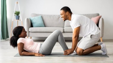 Atletik siyah çift evde birlikte egzersiz yapıyor, karın kasları üzerinde çalışıyor. Neşeli Afro-Amerikalı, spor kıyafetleri içinde bir adam karısının bacaklarını tutarken karın egzersizleri yapıyor, panorama.