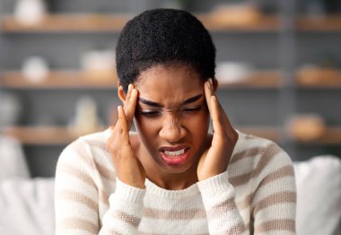 Genç Afro-Amerikalı kadın baş ağrısı ya da migrenden muzdarip, hasta siyah kadın kendini iyi hissetmiyor, akut ağrısı var ve oturma odasında kanepede otururken tapınakları ovuyor, yakın çekim yapıyor.